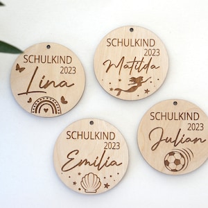 Puede incluir: Cuatro adornos redondos de madera con la palabra "Schulkind" y el a&ntilde;o "2023" grabados en cada uno. Cada adorno tiene un nombre y un dise&ntilde;o diferente: "Lina" con un arco&iacute;ris, "Matilda" con una sirena, "Emilia" con una concha y "Julian" con un bal&oacute;n de f&uacute;tbol.