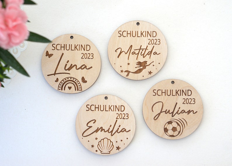 Puede incluir: Cuatro adornos redondos de madera con la palabra "Schulkind" y el a&ntilde;o "2023" grabados en cada uno. Cada adorno tiene un nombre y un dise&ntilde;o diferente: "Lina" con un arco&iacute;ris, "Matilda" con una sirena, "Emilia" con una concha y "Julian" con un bal&oacute;n de f&uacute;tbol.