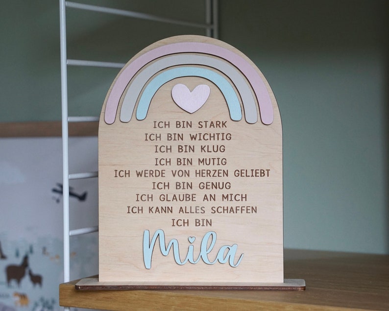 May include: Wooden rainbow-shaped sign with a heart in the center and the text "ICH BIN STARK, ICH BIN WICHTIG, ICH BIN KLUG, ICH BIN MUTIG, ICH WERDE VON HERZEN GELIEBT, ICH BIN GENUG, ICH GLAUBE AN MICH, ICH KANN ALLES SCHAFFEN, ICH BIN Mila".