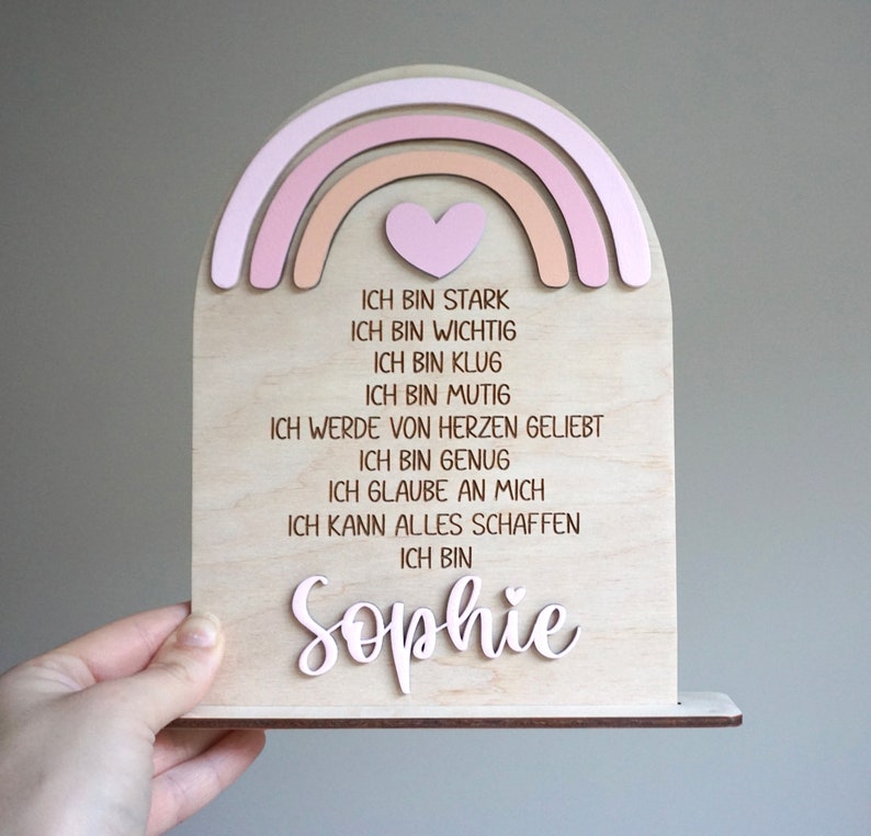 May include: A wooden rainbow with a pink heart and the text "ICH BIN STARK, ICH BIN WICHTIG, ICH BIN KLUG, ICH BIN MUTIG, ICH WERDE VON HERZEN GELIEBT, ICH BIN GENUG, ICH GLAUBE AN MICH, ICH KANN ALLES SCHAFFEN, ICH BIN Sophie"