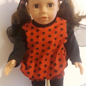 May include: A ladybug costume for a doll, featuring a red dress with black polka dots, black leggings, and black wings. The doll has brown hair and is wearing a black headband with red antennae.