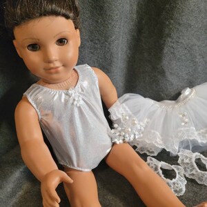 May include: A doll with brown hair and brown eyes wearing a silver leotard. A white tutu with pearl accents and lace trim is next to the doll. The doll is on a gray background.