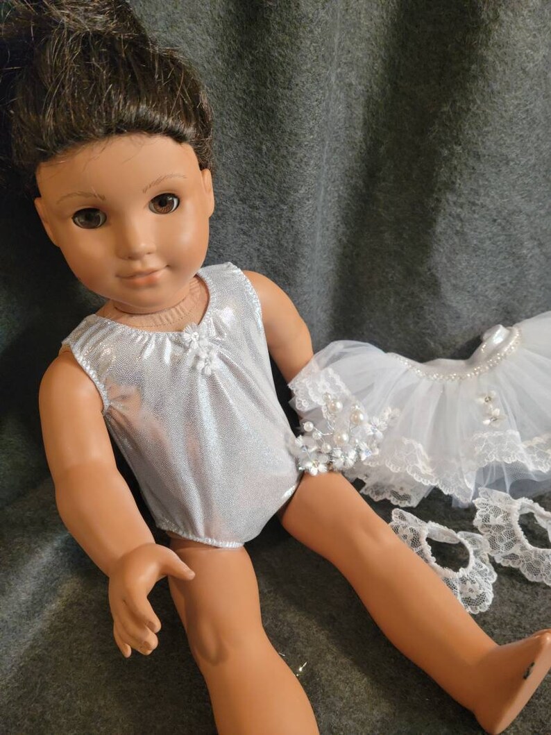 May include: A doll with brown hair and brown eyes wearing a silver leotard. A white tutu with pearl accents and lace trim is next to the doll. The doll is on a gray background.