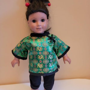 May include: A doll wearing a green and gold Chinese-style top with black pants and a black hair tie with red beads. The doll has brown eyes and brown hair.