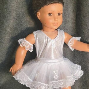 May include: A doll wearing a silver bodice and a white tutu with lace trim. The doll has a small tiara with pearls and flowers in its hair. The doll is wearing lace armbands. The doll is standing on a gray surface.