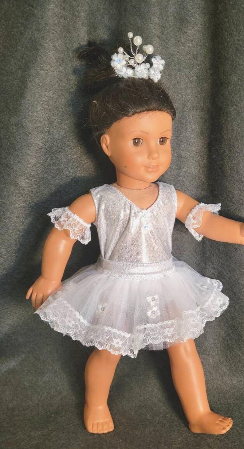 May include: A doll wearing a silver bodice and a white tutu with lace trim. The doll has a small tiara with pearls and flowers in its hair. The doll is wearing lace armbands. The doll is standing on a gray surface.