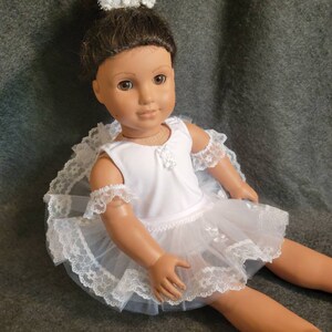 May include: A doll dressed in a white ballerina outfit with a matching crown. The outfit includes a white leotard, a layered tulle skirt, and lace trim. The crown is adorned with pearls and small white flowers.