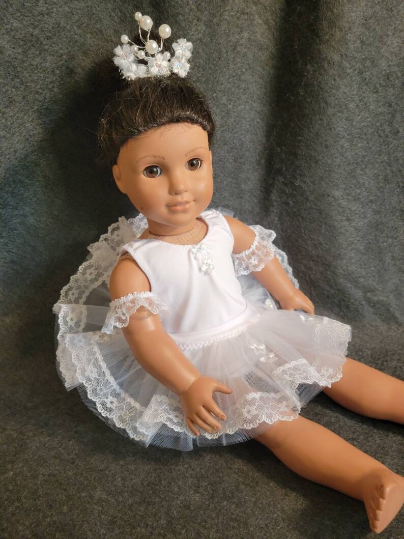 May include: A doll dressed in a white ballerina outfit with a matching crown. The outfit includes a white leotard, a layered tulle skirt, and lace trim. The crown is adorned with pearls and small white flowers.