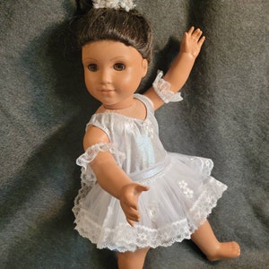 May include: A doll dressed in a white and silver ballet outfit, complete with a tutu and lace trim. The doll has brown hair and is wearing a pearl and crystal tiara. The doll is posed with one arm raised.