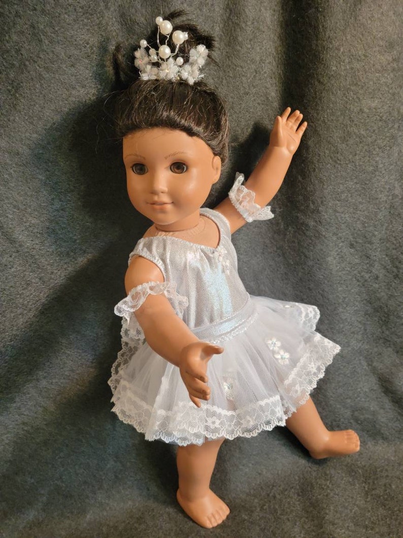 May include: A doll dressed in a white and silver ballet outfit, complete with a tutu and lace trim. The doll has brown hair and is wearing a pearl and crystal tiara. The doll is posed with one arm raised.