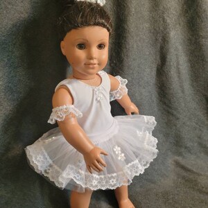 May include: A doll dressed in a white ballerina outfit with a tulle skirt and lace trim. The doll has brown hair and is wearing a crown of white flowers and pearls. The doll is standing on a gray surface.