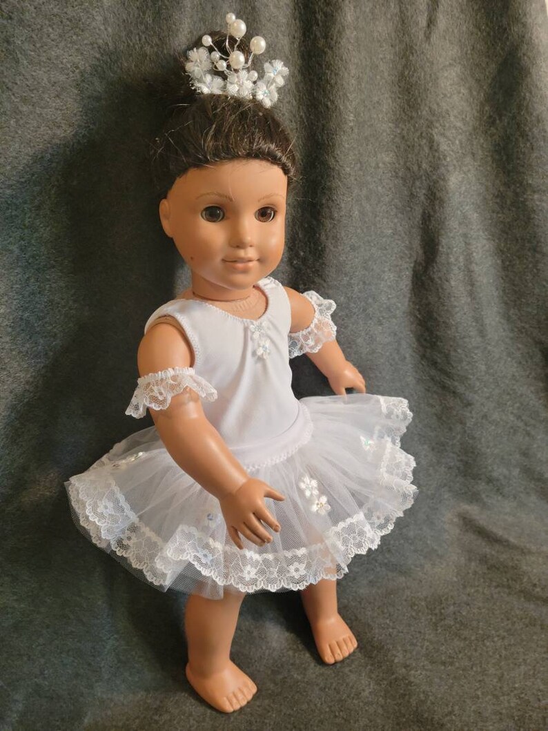 May include: A doll dressed in a white ballerina outfit with a tulle skirt and lace trim. The doll has brown hair and is wearing a crown of white flowers and pearls. The doll is standing on a gray surface.