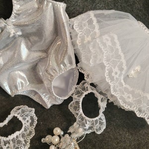 May include: A silver, sparkly bodysuit with matching white lace accessories, including a veil, two lace bands, and a hair comb adorned with pearls. The items are arranged on a dark gray surface.