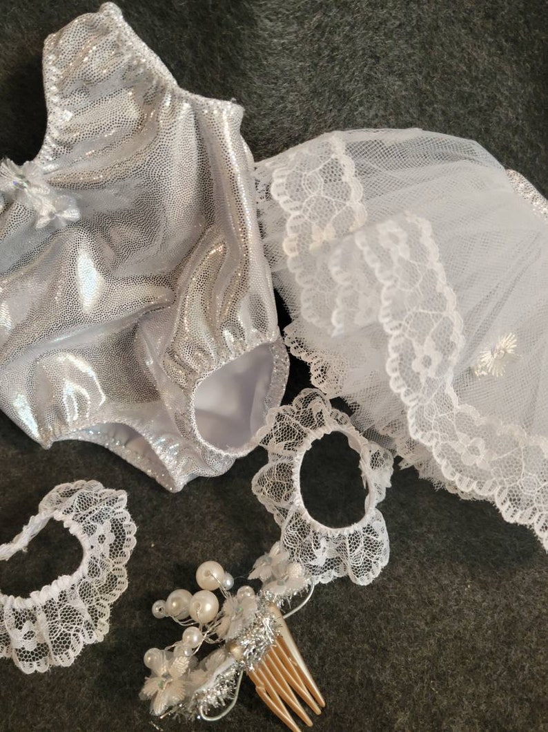 May include: A silver, sparkly bodysuit with matching white lace accessories, including a veil, two lace bands, and a hair comb adorned with pearls. The items are arranged on a dark gray surface.