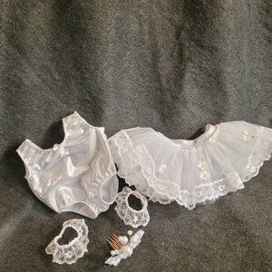 May include: A white baby doll outfit set, including a sparkly bodysuit, a tutu skirt with lace trim, two lace wrist cuffs, and a hair comb with pearls. The set is designed for a doll.