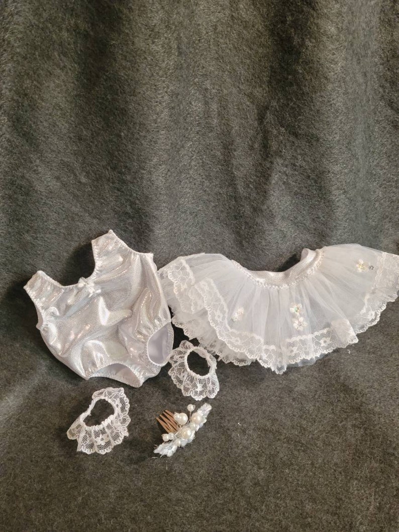 May include: A white baby doll outfit set, including a sparkly bodysuit, a tutu skirt with lace trim, two lace wrist cuffs, and a hair comb with pearls. The set is designed for a doll.
