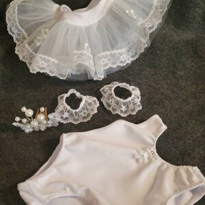 May include: A white doll outfit set, including a tutu skirt with lace trim, two matching lace wrist cuffs, a leotard, and a pearl and floral hair comb. The tutu skirt is made of tulle and lace. The leotard has a floral embellishment.