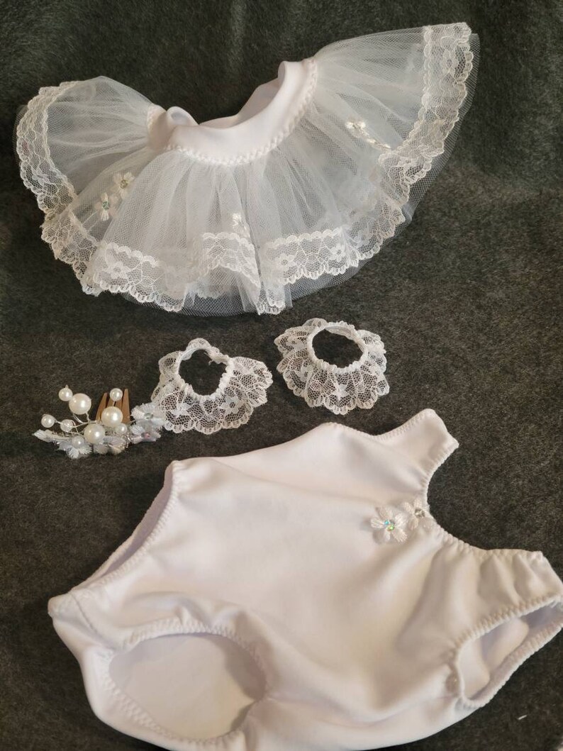 May include: A white doll outfit set, including a tutu skirt with lace trim, two matching lace wrist cuffs, a leotard, and a pearl and floral hair comb. The tutu skirt is made of tulle and lace. The leotard has a floral embellishment.