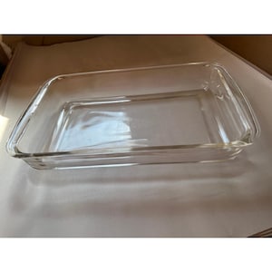 May include: A rectangular clear glass baking dish with rounded corners and integrated side handles. The dish features a smooth, transparent surface and a flat base, designed for oven use, with visible embossed text centered on the bottom interior.
