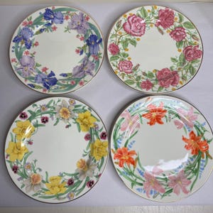 May include: Four white ceramic plates with floral designs. Each plate features a different flower wreath: irises, roses, daffodils, and lilies. The plates have gold rims and are arranged in a square.