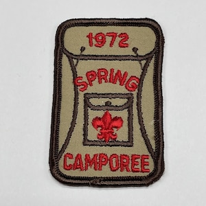 May include: Tan and brown embroidered patch with the text "1972 SPRING CAMPOREE" in red. The patch features a backpack design with a red fleur-de-lis emblem.