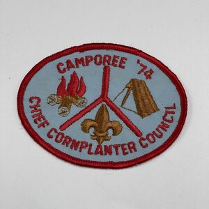 May include: Oval patch with a light blue background and a red border. The patch features the text "Camporee '74" and "Chief Cornplanter Council". It includes embroidered images of a campfire, a tent, and a fleur-de-lis.
