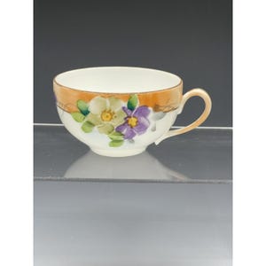 May include: A white teacup with a floral design. The cup has a gold rim and handle, with painted flowers in shades of yellow, purple, and green. The cup is set against a neutral background.
