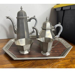May include: A pewter tea service set consisting of a tall coffee pot, a shorter teapot, a creamer, and a sugar bowl, all featuring an octagonal design. The pieces are arranged on a large, octagonal serving tray with a dark wood-grain surface.