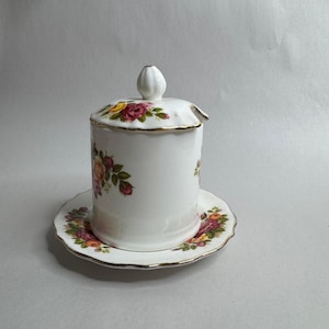 May include: White ceramic sugar bowl with a lid and matching saucer. The bowl and saucer are decorated with a floral pattern of pink, yellow, and red roses with green leaves. The edges are trimmed with gold.