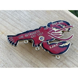 May include: A red and black lobster-shaped pin with gold accents. The pin features the text "THE SETAUKETS EST. 1957" and "DIST 20S C.I.N.Y." The pin has a small blue and gold emblem.