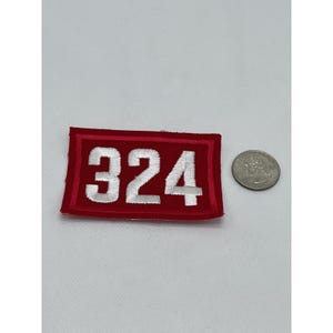 May include: A rectangular red embroidered patch with the number "324" in white thread. The patch has a red border and is shown next to a quarter for size comparison.