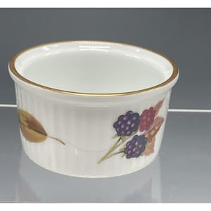 May include: A white ceramic ramekin with a gold rim and a decorative design of blackberries and leaves. The ramekin has a fluted exterior and is suitable for serving individual portions of food.