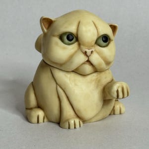 May include: A cream-colored figurine of a cat with green eyes and a grumpy expression. The cat is sitting with one paw raised, and its fur is detailed with subtle shading. The figurine has a rounded shape and a slightly furrowed brow, giving it a unique character.