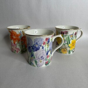 May include: Three white ceramic mugs with floral designs. One mug features orange lilies, another has blue and purple irises, and the third displays yellow daffodils. Each mug has a gold rim and handle, set against a neutral background.