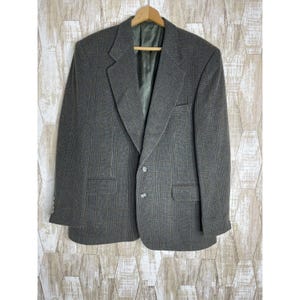 May include: A gray plaid blazer with a two-button closure and notched lapels. The blazer has two front pockets and is hanging on a wooden hanger. The interior lining is a solid green color.