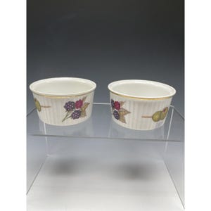 May include: Two white ceramic ramekins with a gold rim and vertical ribbed design. Each ramekin features a painted design of olives, blackberries, and leaves. These ramekins are perfect for serving individual portions of desserts or side dishes.