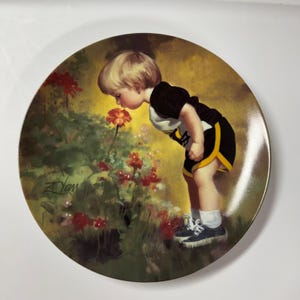 May include: Decorative plate featuring a young child in a black and yellow outfit, smelling a red flower. The plate has a gold rim and a painted scene of a garden with red flowers and green foliage. The artist's signature, "Zdan", is visible.