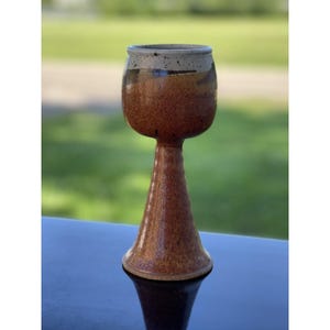 May include: A handmade ceramic goblet with a wide, rounded bowl and a tall, flared stem. The goblet features a speckled brown glaze with a lighter rim and a dark interior. The goblet is displayed on a dark surface with a blurred green background.