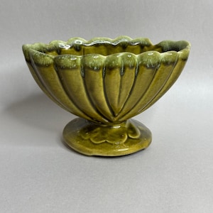 May include: A glossy olive green ceramic bowl with a scalloped rim and vertical fluted detailing. The bowl features a pedestal base and a reactive glaze finish that creates darker green tones along the edges and deep within the fluted grooves.