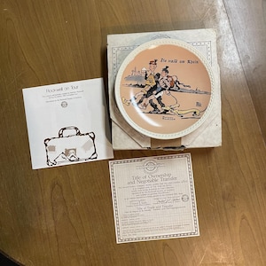May include: A decorative plate with a peach-colored center featuring a Norman Rockwell illustration of people walking. The plate is in its original box with paperwork, including a title of ownership and a brochure titled "Rockwell on Tour."