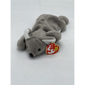 May include: A gray plush koala toy with white accents on the ears and a black nose. The toy has a red heart-shaped tag attached. The koala is lying down on a white surface.