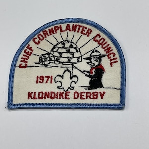 May include: Embroidered patch with a blue border, featuring text "CHIEF CORNPLANTER COUNCIL" and "KLONDIKE DERBY 1971". The patch depicts an igloo, a scout figure, and a fleur-de-lis symbol. The patch is rectangular with a curved top.