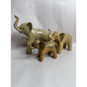 May include: Three decorative elephant figurines in varying sizes. The largest elephant is light beige, the middle one is a lighter beige, and the smallest is a mix of brown and beige. All have trunks raised and ears visible.