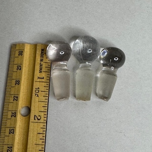 May include: Three clear glass bottle stoppers with spherical tops and frosted, tapered bases. A wooden ruler is positioned to the left, showing the stoppers range from approximately 1.25 to 1.5 inches in height against the imperial scale.