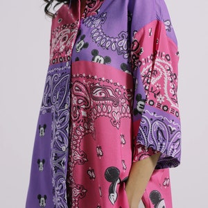 Può includere: Camicia con bottoni con un design patchwork con motivi paisley e sagome di Topolino nei toni del rosa e del viola. La camicia ha un colletto e maniche a tre quarti.