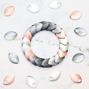 May include: A silver, gray, and rose gold dragon scale bracelet made of metal. The bracelet is made up of individual scales that are linked together. The scales are arranged in a circular pattern, with the rose gold scales in the center and the silver and gray scales on the outside. The bracelet is surrounded by individual scales in the same colors.