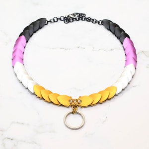 May include: A black, white, pink, and gold chainmail necklace with a silver ring attached. The necklace is made of small, overlapping metal scales.