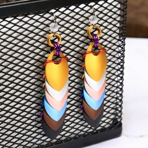 May include: A pair of dangle earrings with a heart-shaped design. The earrings are made of metal and have a rainbow colour scheme. The earrings are hanging from a black wire mesh background.
