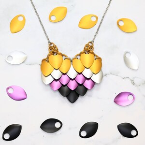 May include: A necklace with a heart-shaped pendant made of metal scales in gold, silver, pink, and black. The scales are arranged in a pattern that resembles a dragon's scales. The necklace is surrounded by loose metal scales in gold, silver, pink, and black.