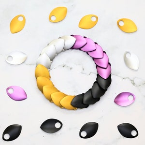 Könnte beinhalten: Ein Satz aus Metallschuppen in verschiedenen Farben, darunter Gold, Silber, Schwarz, Rosa und Lila. Die Schuppen sind in einem Kreis angeordnet, wobei die goldenen Schuppen oben und die schwarzen Schuppen unten sind. Die Schuppen sind alle gleich groß und gleich geformt und haben ein Loch in der Mitte. Die Schuppen werden verwendet, um Schmuck oder andere Handarbeiten herzustellen.